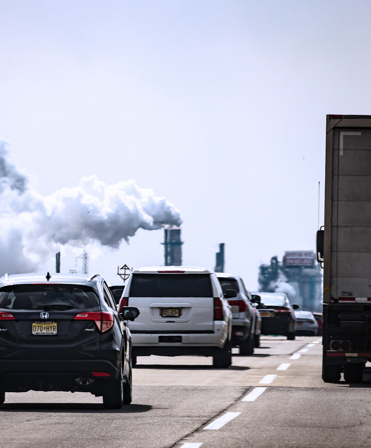 Smoggy view of cars and trucks on highway, with smoke billowing from factory in the background.